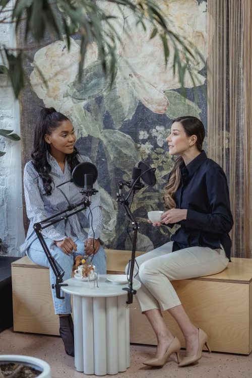 Zwei Frauen sitzen sich bei einem Podcast-Gespräch auf einer Holzbank gegenüber, zwischen ihnen zwei Mikrofone.