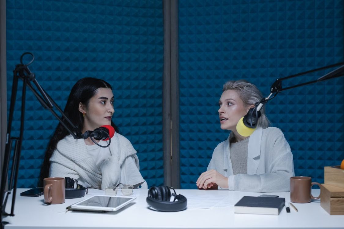 Zwei Frauen nehmen eine Podcast-Folge in einem Studio mit zwei Mikrofonen und dunkelblauen Akustikwänden auf.